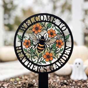 May include: A circular black metal garden stake featuring a cutout design of a bumblebee surrounded by orange flowers and green leaves. The text WELCOME TO HEATHER'S GARDEN is integrated into the circular frame surrounding the central bee motif.