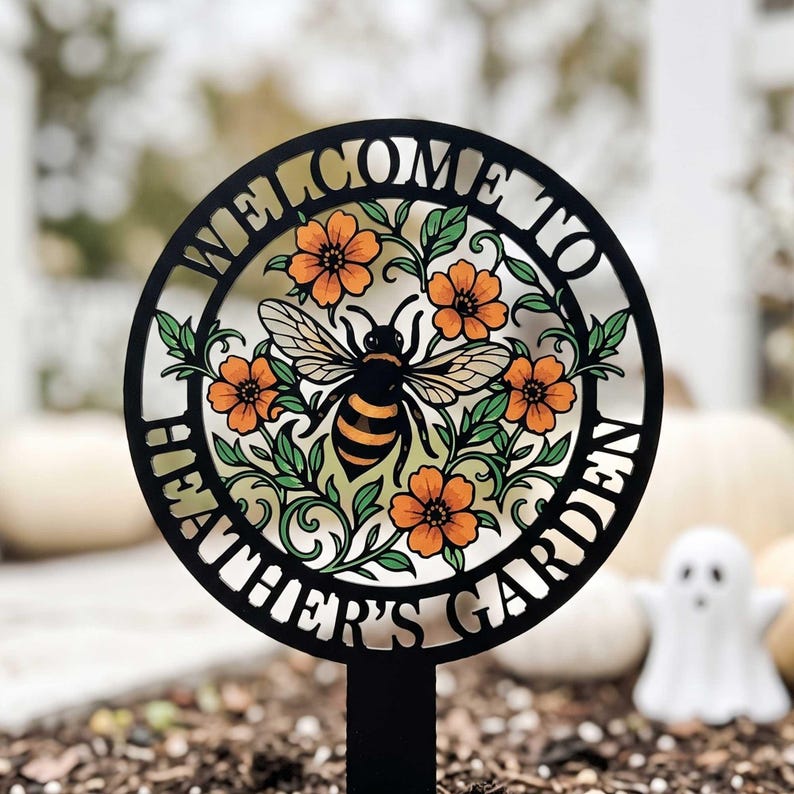 May include: A circular black metal garden stake featuring a cutout design of a bumblebee surrounded by orange flowers and green leaves. The text WELCOME TO HEATHER'S GARDEN is integrated into the circular frame surrounding the central bee motif.