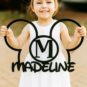 May include: A black wooden sign in the shape of Mickey Mouse ears, with the letter "M" inside a circle and the name "Madeline" below. The sign is held by a smiling child wearing a white dress.