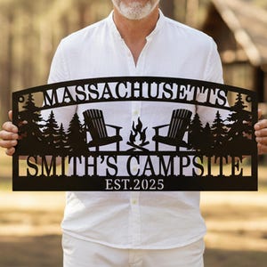 May include: Black metal sign with the text "MASSACHUSETTS" above "SMITH'S CAMPSITE EST. 2025". The sign features silhouettes of trees, Adirondack chairs, and a campfire. The sign is held by a person.
