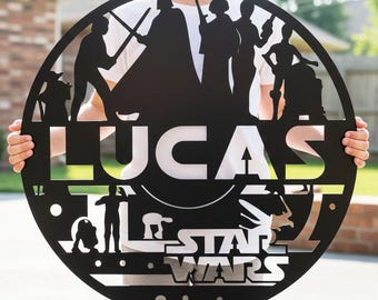 May include: Black metal Star Wars themed sign with the name "LUCAS" cut out. The sign features silhouettes of characters and the Star Wars logo. The sign is round and is being held up by a person.