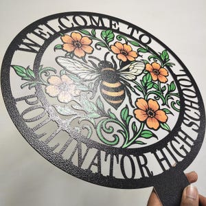 May include: A circular black sign with a textured finish featuring the text "WELCOME TO POLLINATOR HIGH SCHOOL" cut out along the border. The center displays a detailed illustration of a bee surrounded by orange flowers and green leaves.