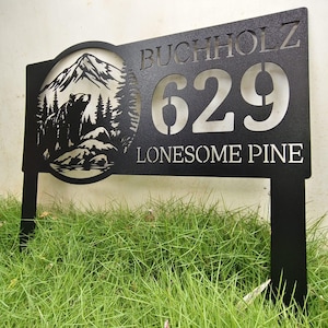 Peut inclure: Panneau d'adresse en métal noir avec le nom "BUCHHOLZ", le numéro "629" et les mots "LONESOME PINE". Le panneau présente un motif découpé d'un ours, de montagnes et d'arbres. Le panneau est monté sur deux poteaux et planté dans l'herbe.