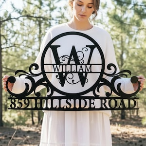 May include: Black metal house sign with the name "WILLIAM" and address "859 HILLSIDE ROAD". The sign features a large letter "W" in a circular design with decorative scrollwork. The sign is held by a person in a white sweater.