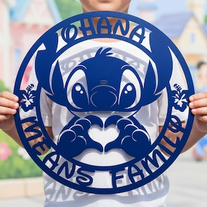 Op de afbeelding: Een blauw metalen wandkunstwerk met het personage Stitch uit Lilo & Stitch. Het ontwerp is cirkelvormig, met de woorden "OHANA MEANS FAMILY" uitgesneden rond de afbeelding van het personage. Stitch maakt een hartvorm met zijn handen.