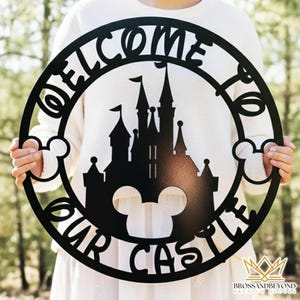 Puede incluir: Letrero circular de metal negro con las palabras "WELCOME OUR CASTLE" y una silueta de un castillo. El letrero tiene formas de orejas de Mickey Mouse en los lados. El letrero se sostiene frente a un fondo verde borroso.