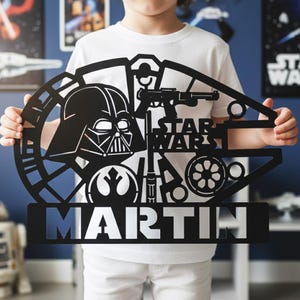 Może przedstawiać: Czarna metalowa grafika ścienna o tematyce Gwiezdnych Wojen przedstawiająca Dartha Vadera, blaster i logo Gwiezdnych Wojen. Imię "MARTIN" jest wyświetlane pogrubioną czcionką pod obrazami Gwiezdnych Wojen. Grafika jest trzymana przez osobę ubraną w białą koszulkę.