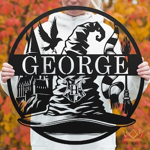 May include: Black metal circular sign with the name "GEORGE" in white. The design includes a sorting hat, a raven, a scarf, a broom, and a crest. The background is blurred with fall foliage.
