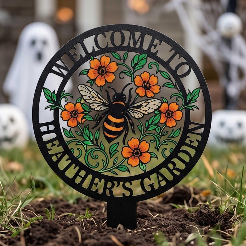 May include: A circular black metal garden stake featuring a cutout design of a bee surrounded by orange flowers and green leaves. The text "WELCOME TO HEATHER'S GARDEN" is integrated into the circular frame, creating a decorative outdoor sign.