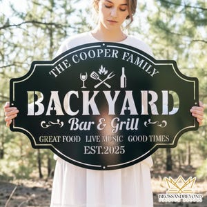 May include: A black metal sign with a decorative border, featuring the text "THE COOPER FAMILY BACKYARD Bar & Grill" in silver lettering. The sign includes icons of a wine glass, grill, and bottle, with the words "GREAT FOOD LIVE MUSIC GOOD TIMES EST.2025".