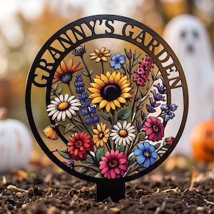 Puede incluir: Un estaca de jardín circular de metal negro con el texto "GRANNY'S GARDEN" recortado en el borde superior. El centro muestra una ilustración colorida de varias flores, incluyendo un gran girasol amarillo, margaritas blancas y lavanda.