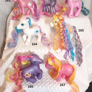 Può includere: Collezione di sette pony giocattolo colorati. I pony sono in varie tonalità di rosa, viola e bianco, con lunghe criniere e code multicolori. Ogni pony ha segni e disegni unici, tra cui fiori, arcobaleni e cuori.
