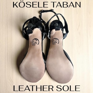 K&ouml;nnte beinhalten: Paar schwarze High-Heel-Sandalen mit Kn&ouml;chelriemen und gl&auml;nzendem Finish. Die Sohlen sind beige mit einem aufgedruckten Design. Der Text "K&Ouml;SELE TABAN" und "LEATHER SOLE" ist zu sehen.