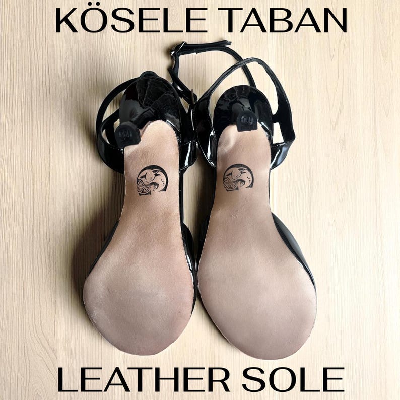 K&ouml;nnte beinhalten: Paar schwarze High-Heel-Sandalen mit Kn&ouml;chelriemen und gl&auml;nzendem Finish. Die Sohlen sind beige mit einem aufgedruckten Design. Der Text "K&Ouml;SELE TABAN" und "LEATHER SOLE" ist zu sehen.