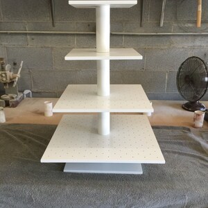 May include: White four-tiered display stand with a white base and a white pole connecting the tiers. Each tier has a white surface with small holes.