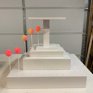 May include: A tiered, white cake stand with a rectangular top and base, and a central support. Several cake pops in shades of pink, orange, and yellow are placed on the stand. The cake pops are on wooden sticks.