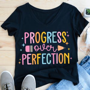 May include: Black v-neck t-shirt with the text "PROGRESS over PERFECTION" in colorful letters. The design includes a pencil and stars. The shirt is paired with gray sneakers and blue jeans.