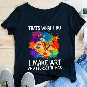 以下が含まれることがあります： 「THAT'S WHAT I DO I MAKE ART AND I FORGET THINGS」のテキストが入った黒いVネックTシャツ。鮮やかなマルチカラーのペイントスプラッターの背景に、3本の筆とカラフルなパレットが描かれています。