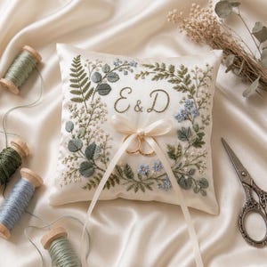 May include: Ivory satin ring pillow with embroidered floral design in green and blue. Features the initials "E & D" and a cream-colored ribbon holding two gold wedding rings. Spools of thread and scissors are in the background.