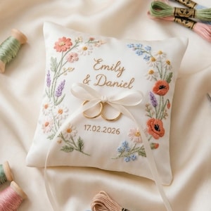 Peut inclure: Coussin d'alliances brodé avec un motif de couronne florale, avec les noms "Emily & Daniel" et la date 17.02.2026. Comprend deux alliances dorées, entourées de bobines de fil, de ciseaux et d'un dé à coudre. Le coussin est blanc avec des broderies colorées.