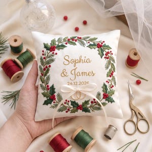 Pode incluir: Uma almofada quadrada branca para alianças com uma coroa de azevinho bordada em verde com bagas vermelhas. O centro apresenta o texto bordado a dourado Sophia & James e a data 24.12.2026. Uma fita creme segura dois anéis dourados.