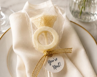 May include: A ring-shaped lollipop with a glittery diamond-shaped top, wrapped in clear plastic. A gold ribbon ties a small white circular tag to the stick, featuring the black printed text "You're Next!" The lollipop rests on a folded cream napkin.
