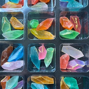 May include: A clear plastic container filled with colorful, translucent, irregularly shaped candy pieces. The candy is arranged in rows and columns, with each compartment containing a different color.