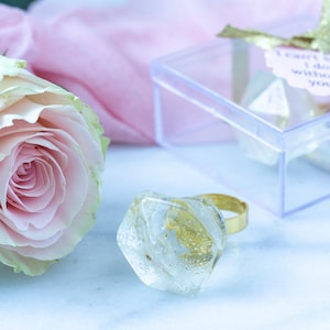 May include: A clear glass ring with gold accents and a delicate design. The ring is sitting on a white surface with a pink fabric and a clear box in the background.