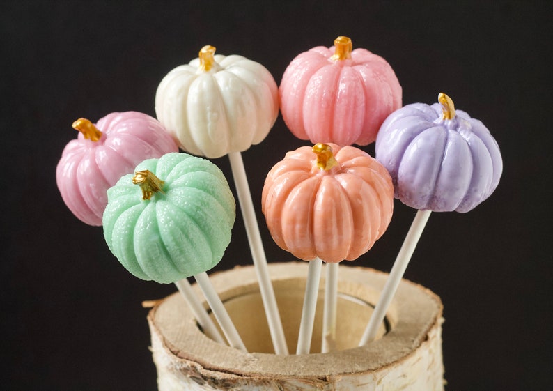 Puede incluir: Seis piruletas con forma de calabaza de colores pastel en palos, dispuestas en un soporte de tronco de abedul. Las piruletas son blancas, rosas, lavanda, melocot&oacute;n y verdes.