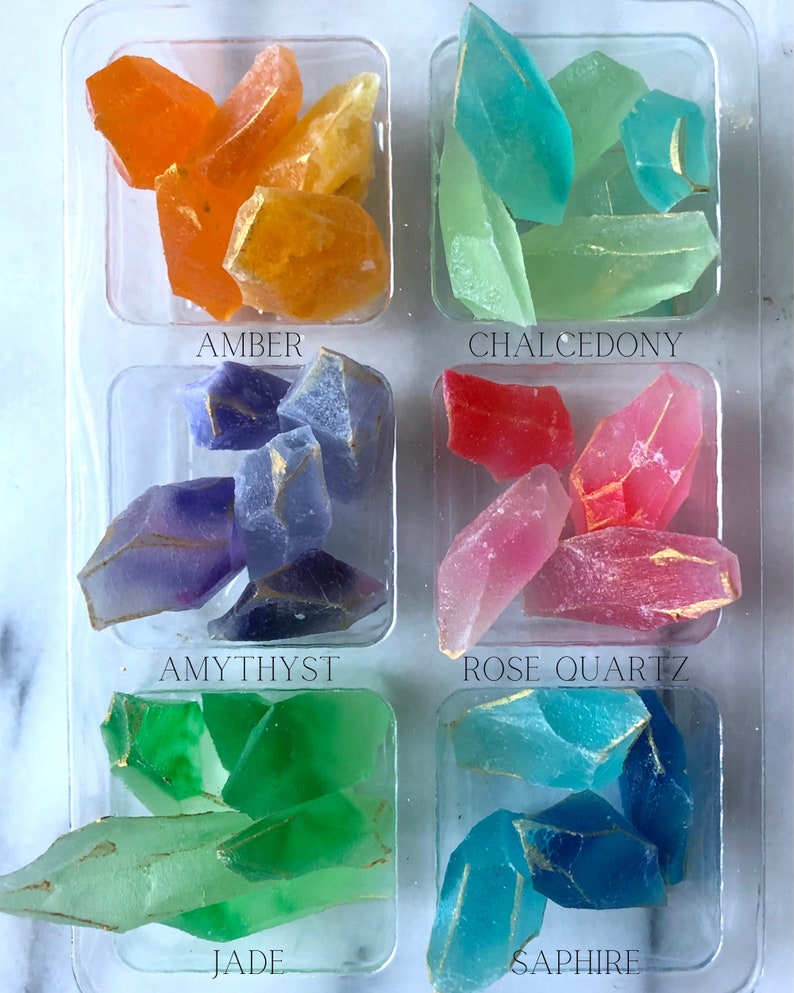 May include: A close-up of a tray of six different colored crystal-shaped soaps. The soaps are arranged in two rows of three. The top row from left to right is amber, chalcedony, and rose quartz. The bottom row from left to right is amethyst, jade, and sapphire.