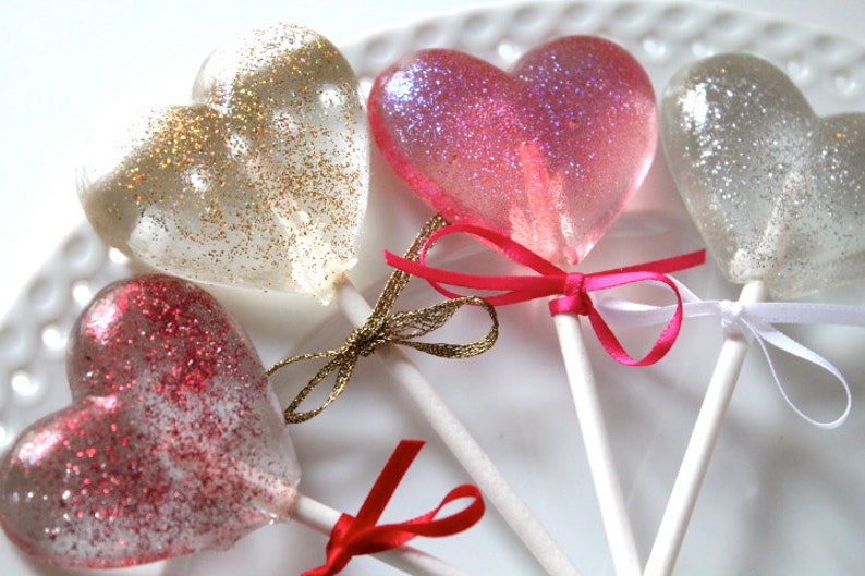 May include: Four heart-shaped lollipops with glitter on a white surface. The lollipops are tied with ribbons in gold, pink, and white. The lollipops are clear with glitter in gold, pink, and silver.