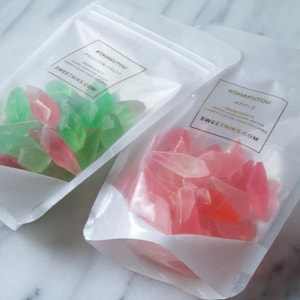 May include: Two clear plastic bags filled with colorful candy. The bag on the left contains green and pink candy. The bag on the right contains pink candy. The bags have labels that say "Kohakuto" and "Sweetniks.com".