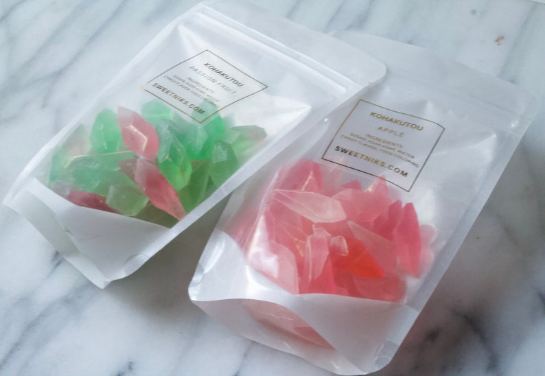 May include: Two clear plastic bags filled with colorful candy. The bag on the left contains green and pink candy. The bag on the right contains pink candy. The bags have labels that say "Kohakuto" and "Sweetniks.com".