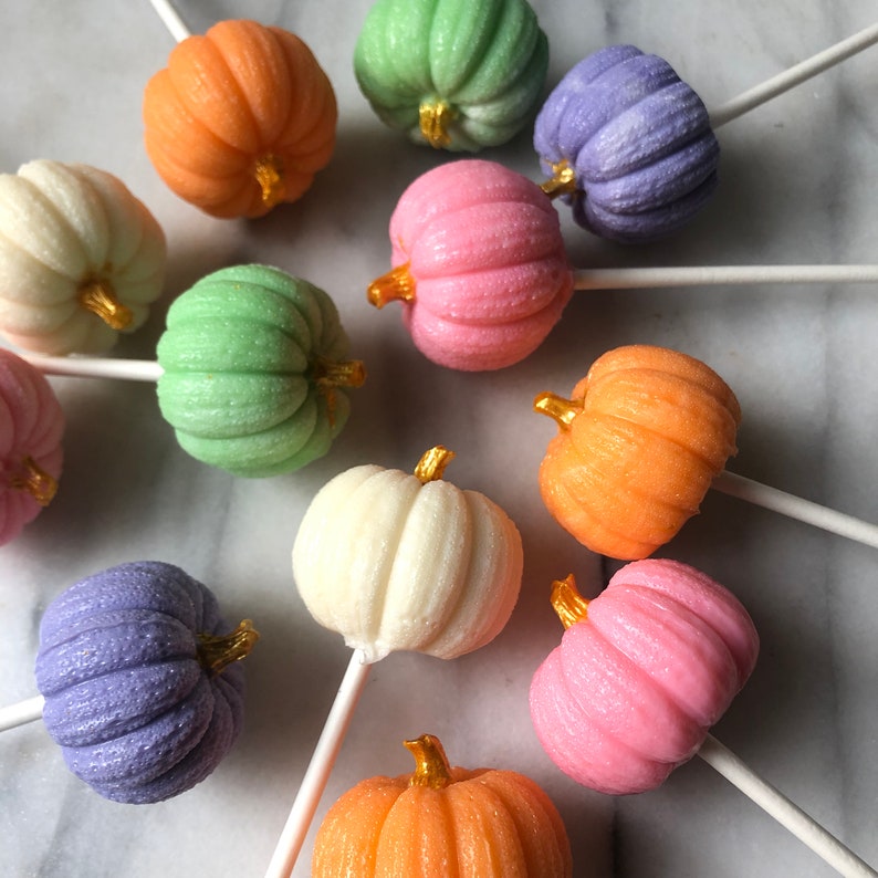 Puede incluir: Surtido de piruletas con forma de calabaza de colores pastel en palos. Las piruletas son en tonos de rosa, verde, morado, naranja y blanco.