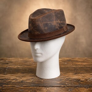 Op de afbeelding: Een bruine fedora hoed met een verweerde, doorleefde textuur en een donkerbruine suède band rond de basis. De hoed heeft een ingedeukte kroon en een gestructureerde rand, met een gevlekt patroon over het gehele oppervlak van het materiaal.