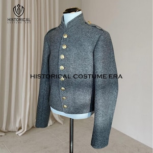 Op de afbeelding: Een grijze jas in militaire stijl met een wolachtige textuur, een hoge kraag, lange mouwen en een verticale rij van zeven gepolijste goudkleurige knopen aan de voorzijde. Het kledingstuk staat op een paspop en bevat de tekst HISTORICAL COSTUME ERA.