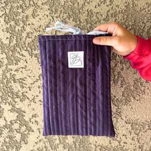 May include: A purple and white striped fabric bag with a light blue drawstring closure. A white square label with a black abstract design is centered on the bag. The bag is held by a person wearing a red long-sleeve shirt.