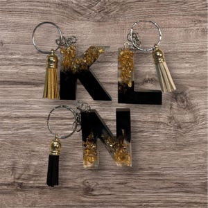 Può includere: Portachiavi con le lettere K, L e N. Ogni lettera è nera con dettagli in foglia d'oro e un nappa abbinata. I portachiavi hanno anelli argentati.