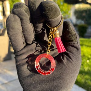 Può includere: Un portachiavi fatto a mano con un cerchio di resina trasparente con glitter rossi a forma di cuore. Ha una catena color oro, un nappa rosa e una chiusura color oro. Lo sfondo è sfocato, con vegetazione e luce solare.