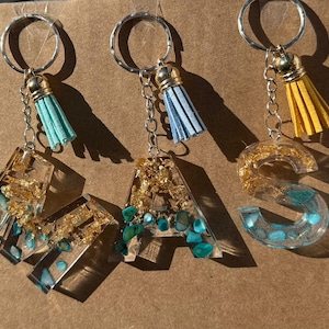 Può includere: Tre portachiavi con ciondoli a forma di lettera in resina trasparente. Le lettere sono riempite di scaglie d'oro e pietre turchesi. Ogni portachiavi ha un anello argentato, un nappa e una catena. Le nappe sono verde acqua, azzurre e gialle.