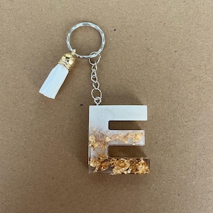 Può includere: Un portachiavi con la lettera "E" in resina trasparente con inserti in foglia d'oro. Il portachiavi ha una catena argentata, un portachiavi argentato e un nappa bianca con cappuccio dorato.