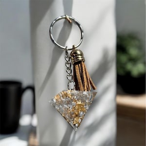 Può includere: Un portachiavi argentato con un ciondolo a forma di diamante. Il ciondolo è trasparente con accenti in foglia d'oro. Un nappa marrone e una catena d'argento pendono dall'anello del portachiavi.