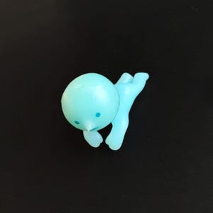 以下が含まれることがあります： ダイビングや水泳のポーズをとった、デフォルメされた人型の小さな水色のフィギュア。滑らかで丸い頭部には、目として2つの小さな濃い青色の点があり、鼻先は尖った形状をしています。手足は外側に伸びています。