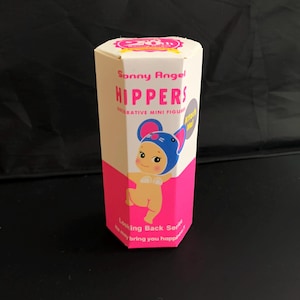 May include: A hexagonal white and pink cardboard box for a Sonny Angel Hippers decorative mini figure from the Looking Back Series. The packaging features a graphic of a character wearing a blue mouse hat and the text Attach Me! in a grey circle.