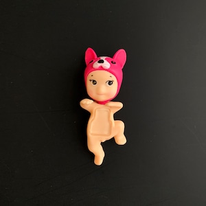 May include: A small figurine of a baby wearing a bright pink animal-themed hood with white accents. The figure has a light peach-coloured body with a recessed rectangular area on the chest, depicted in a dynamic pose with one arm raised and one leg bent.