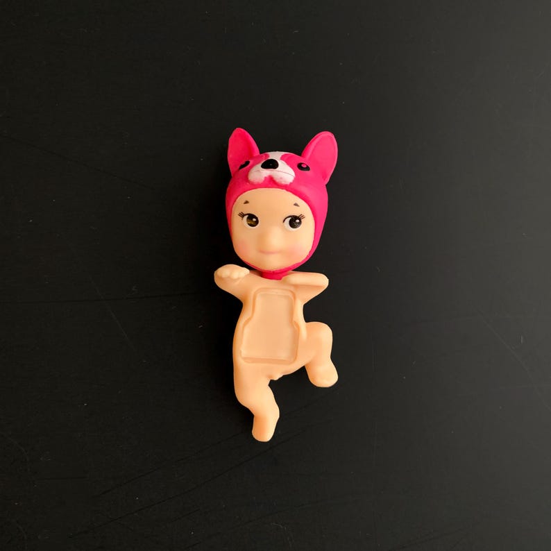 May include: A small figurine of a baby wearing a bright pink animal-themed hood with white accents. The figure has a light peach-coloured body with a recessed rectangular area on the chest, depicted in a dynamic pose with one arm raised and one leg bent.