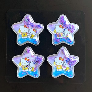May include: Four star-shaped shaker charms featuring a Hello Kitty graphic with a yellow outfit and a red bow. Each clear plastic charm contains a liquid-filled center with loose purple star-shaped glitter and small white beads inside.