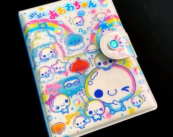 Könnte beinhalten: Ein weißes Notizbuch mit Knopfverschluss. Der Einband zeigt bunte Cartoonfiguren, Regenbogen und japanischen Text. Das Design umfasst blaue, rosa, gelbe und grüne Akzente.