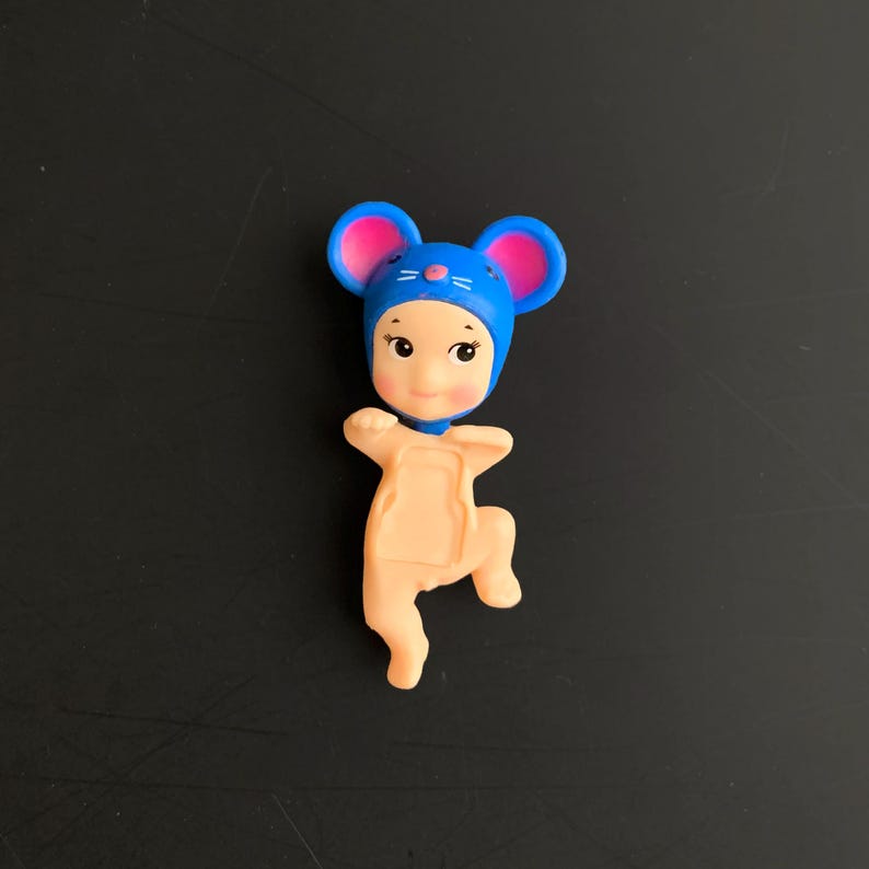 May include: A small figurine featuring a character wearing a blue mouse hood with pink inner ears. The character has a peach-coloured body, a rounded face with dark eyes, and a small nose. The figure is posed with arms and legs bent in a dynamic stance.