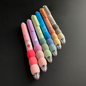 May include: A collection of colorful pens with a unique design. Each pen features a different pastel color, a textured grip, and a clear tip. Some pens have small illustrations and text, including the word "HUG".
